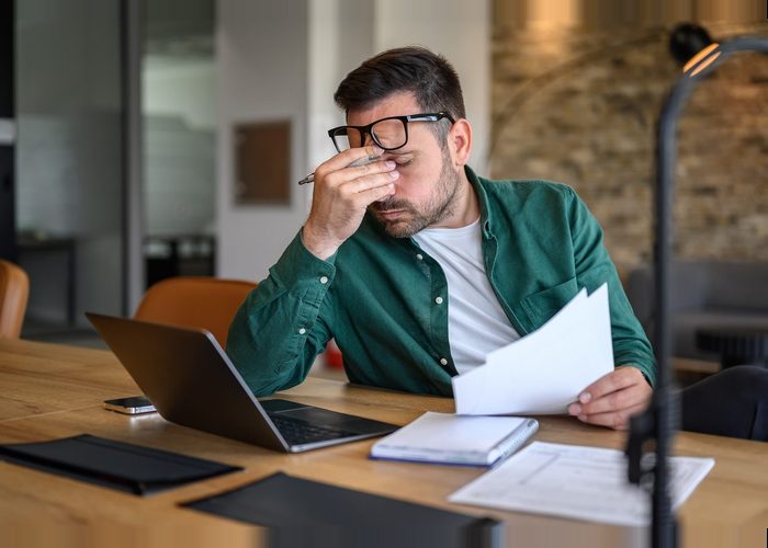 stressed male financial advisor rubbing eyes while working over laptop and analyzing reports at desk